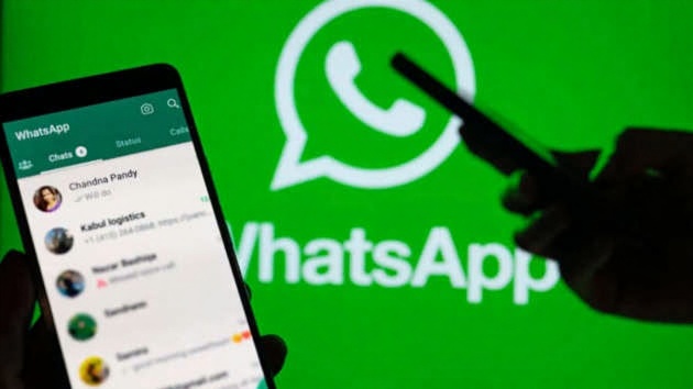 Hands holding a smartphone with the WhatsApp chat list open, while another phone in the background takes a picture of the large, glowing WhatsApp logo on a green screen.